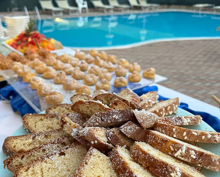 media.Slide_Buffet di frutta e dolci hotel Abarth, dettaglio ciambella in primo piano e piscina sullo sfondo